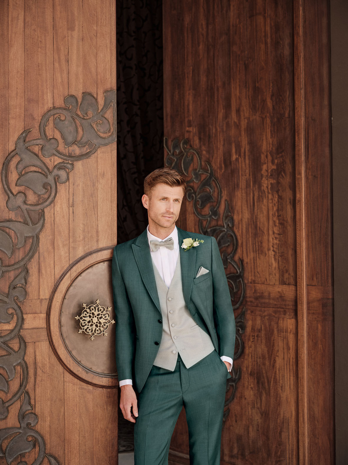 Stilvoll heiraten im Wilvorst-Anzug – Eleganz in modernem Grün Bräutigam in grünem Wilvorst-Anzug mit hellgrauer Weste, Fliege und Ansteckblume vor kunstvoller Holztür – moderne Hochzeitsmode für Herren
